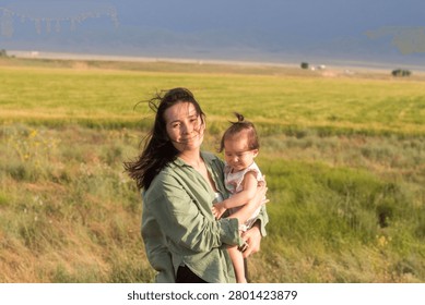 A horizontal, eye-level medium shot capturing a tender moment of motherhood in a rural landscape. A young Central Asian woman with long dark hair gently blown by the wind smiles warmly at the camera while holding her young toddler daughter securely in her arms. The mother is dressed in a casual, loose-fit sage green button-up shirt over a white top, while the baby girl wears a light floral sleeveless summer top with her hair tied in a small top knot. They are standing in a vast, vibrant green and yellowish grass field under soft evening sunlight. In the far distance, a soft horizon line meets a dramatic, dark overcast sky, creating a beautiful contrast between the warm, bright field and the moody background elements.