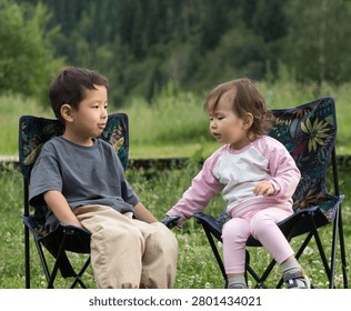 A horizontal, eye-level medium shot of two young children sitting next to each other in dark blue folding camp chairs with a colorful tropical leaf pattern. On the left, a young boy of Central Asian ethnicity is dressed in an oversized dark grey t-shirt and loose beige trousers, sitting calmly and looking toward his companion. On the right, a smaller toddler girl with fine brown hair is wearing a pink and white raglan long-sleeve top, light pink leggings, socks, and sneakers. She leans slightly toward the boy, gesturing or talking to him. They are positioned outdoors on a green grass lawn or meadow with a soft-focus background of lush green trees and a forested hill, capturing a candid, peaceful moment of childhood friendship and outdoor recreation.