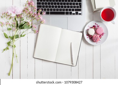 Horizontal flat top view of white wooden table with grey metal laptop with keyboard open, flowers with pink buds, macarons and tea, copybook with blank sheets lying open ready for taking notes