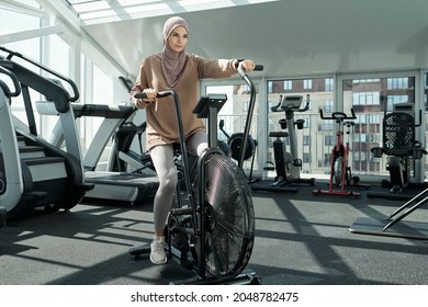 Horizontal full body shot of active young adult Muslim woman wearing hijab exercising in gym using stationary bike machine - Powered by Shutterstock