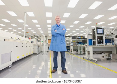 Horizontal Full Length Shot Of An Engineer Standing With Crossed Arms In A High Tech Electronic Factory Smiling At The Camera.