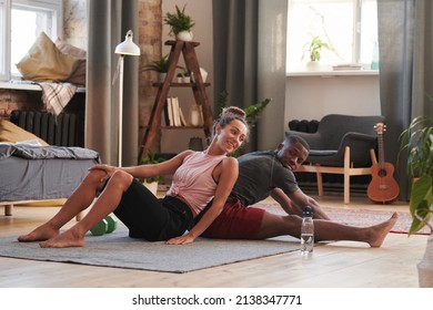 Horizontal full shot of lovely young woman sitting on floor leaning onto her African American boyfriends back helping him with stretching exercise - Powered by Shutterstock