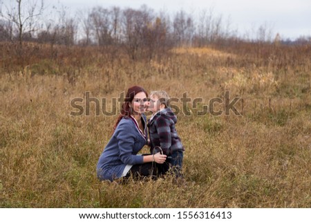 Horizontal full-length portrait of handsome little boy kissing his beautiful young mother crouching next to him in field during a grey fall afternoon