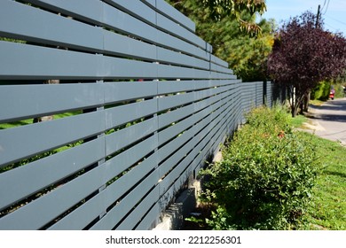 Horizontal Gray Metal Fence In Diminishing Perspective. Property Safety And Home Ownership Concept. Village Style Street Scene. Light Gray Paint. Lush Green Shrub And Grass Along The Asphalt Road. 