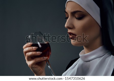 Horizontal half-turn close-up portrait of a nun, posing on a black background. She's wearing dark nun's clothing. The nun holding a glass of wine in a right hand and looking at it.  