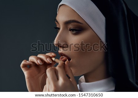 Horizontal half-turn close-up portrait of a nun, posing on a black background. She wearing dark nun's clothing. The nun brings a cigarette to her mouth and licking it.  
