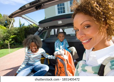 Horizontal head and shoulder profile shot of a joyous mother standing by two children sitting in boot of a car with luggage smiles at the camera.