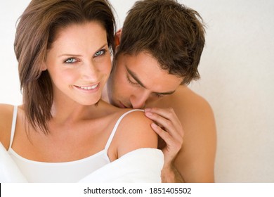 Horizontal Head And Shoulder Shot Of A Young Couple With The Man Kissing On Woman's Shoulder From Behind As She Smiles At The Camera.