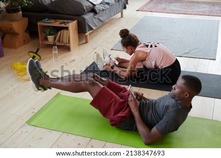 Horizontal high angle view shot of young Black man and his Caucasian girlfriend doing morning workout at home and watching something on smartphones