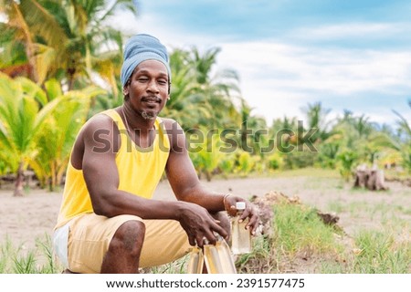 Horizontal image of an African American man sitting on a tree trunk on the beach. Black man with slanted eyes wearing a yellow sleeveless shirt and shorts with green beach palms in the background.