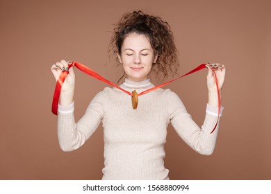 Horizontal Image Of Beautiful Curly Haired Woman Stands Against Beige Wall Background With Closed Eyes, Wears Brown Casual Cashmere Jumper, Decorating Study Room With Ginger Cookies On Christmas Eve.