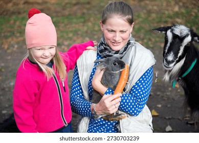 Horizontal image capturing a smiling middle aged woman and a young girl feeding a rabbit with an orange carrot as a goat looks on. Warm colors emphasize connection and curiosity.