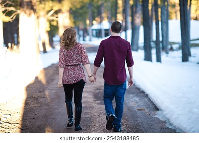 Horizontal image of couple walking down a dirt road together and holding hands. There is snow and trees lining the dirt road. 