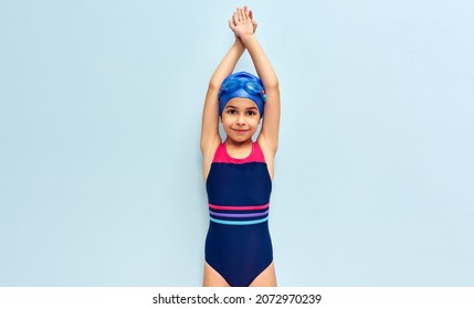 Horizontal Image Of A Cute Little Girl In Goggles, A Swimsuit, And A Blue Swimming Cap Get Ready Before Diving Isolated Blue Studio Background. Kid In Swimsuit Making Swimming Gesture Before The Dive.