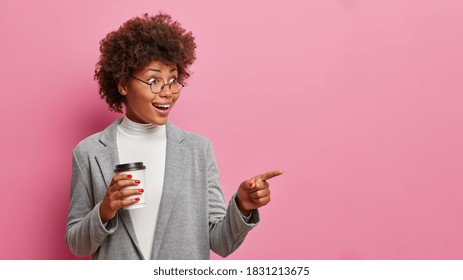 Horizontal image of happy surprised business lady with glad reaction indicates index finger away into distance poses against rosy background drinks take away coffee dressed in formal clothing - Shutterstock ID 1831213675