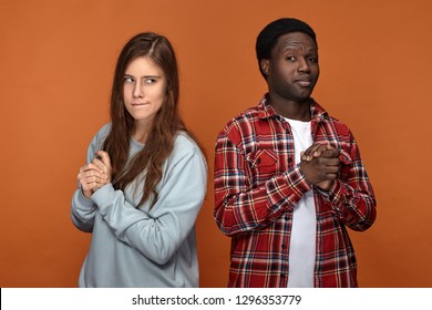 Horizontal Image Of Interracial Couple Pulling Tricks On Each Other, Posing Isolated Against Blank Orange Studio Wall Background, Looking Suspicious, Planning Trick Or Prank, Rubbing Hands