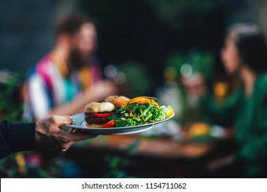 Horizontal Image Of Male Hand Waiter Serving Plate Of Vegetarian Burger Dinner In Restaurant To A Young Couple.