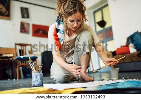 Horizontal image of a pretty female artist sitting on the floor in the art studio and painting on paper with a brush. A woman painter with glasses painting with watercolors in the workshop.