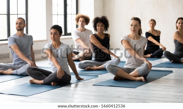 Horizontal image seven multi-ethnic people during\
yoga class seated in lotus position performing Easy Twist Pose or\
Parivrtta Sukhasana. Exercise decrease feeling of nervousness,\
reduce stress\
concept
