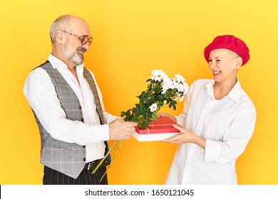 Horizontal Image Of Shy Awkward Grandfather Wuth Gray Beard Holding Flowers And Box Of Present Congratulating His Mature Girlfriend On Her Birthday. Cute Happy Elderly Couple On First Date