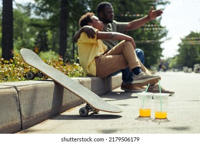 Horizontal Image Of Skateboard And Cold Drinks Standing On The Ground With Dad And Son Resting Together In Background