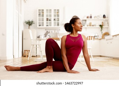 Horizontal image of sporty stylish young Afro American woman in sports clothes practicing yoga, sitting on mat with one knee bent, turning head. Healthy lifestyle, wellbeing and activity concept