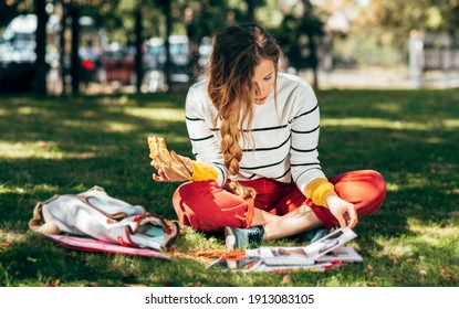 Horizontal Image Of A Student Female Sitting On The Green Grass At The College Campus On A Sunny Day, Have Lunch And Studying. The Hungry Young Woman Takes A Rest Learning And Eating In The Park.