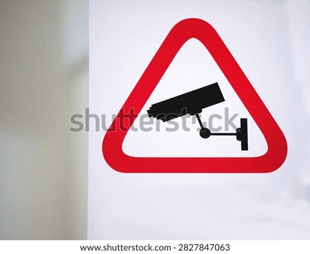 Horizontal image of surveillance camera warning sign with red triangle on light background, highlighting security monitoring and safety awareness concept.