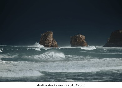 Horizontal image of two large rocks battered by the waves of the sea raging in the storm, the waves crashing against the rocks under a dark sky.