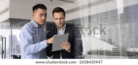 Horizontal image of two men working together using tablet in modern city highrise building office, researching, review new AI platform or application, analyzing business performance or market trends