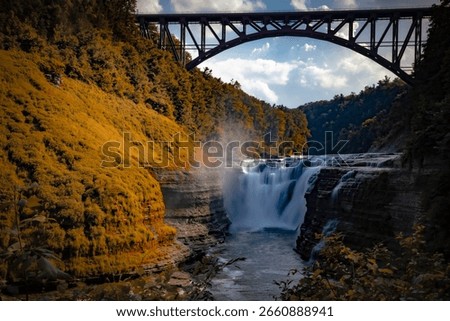 horizontal image of the Upper Falls, under a bridge,  surrounded by golden trees, in Letchworth State Park in New York.