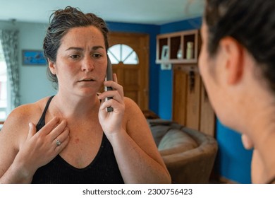 Horizontal image of young female in mirror reflection calling doctor about concerns of skin cancer on chest.