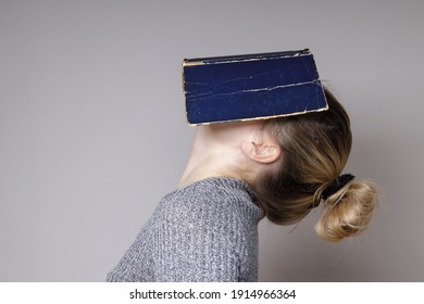 Horizontal image of a young woman covering her face with a book. A beautiful woman in a gray knitted sweater, hiding her face behind a blue book cover, with her head thrown back. Book concept