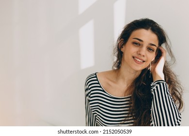 Horizontal indoor portrait of charming charismatic photogenic Caucasian female with dark long wavy hair posing against white wall with sun rays and copy space for your advertising content