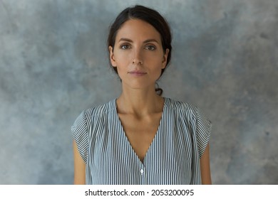 Horizontal indoor portrait of charming young lady posing for ID or passport photo, having calm face expression, looking straight at camera without smile, standing against textured studio wall
