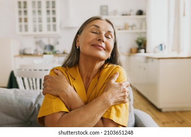 Horizontal indoor portrait of happy elderly woman hugging herself, giving support, showing self-respect and love, sitting on couch with closed eyes in yellow shirt. Human emotions, body language