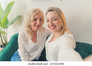 Horizontal Indoor Shot In An Apartment Of Two Blonde Well-dressed Attractive Millenial Female Siblings Sitting On A Dark-green Couch. High Quality Photo