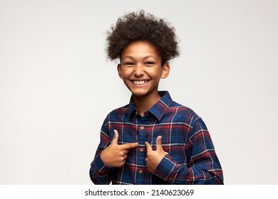 Horizontal joyful little African American man of 12 pointing at himself with cheerful toothy smile after winning prize, posing against white studio wall with copy space for your advertising content