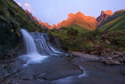 A Horizontal Landscape Photograph Of A Beautiful Waterfall In Front Of The Drakensberg Escarpment Mountains At Sunrise With Beautiful Golden Sunrise Light On The Cliffs In The Distance.