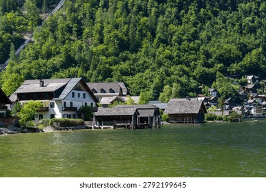A horizontal landscape view of authentic alpine residential houses and historic wooden boathouses built directly over the clear water of a mountain lake in Austria. The scene highlights traditional weathered wooden roofs, cozy lakeside piers, and a steep, dense green forest covering the background hillside on a bright summer day. Great for European travel, alpine lifestyle, and vacation concepts.