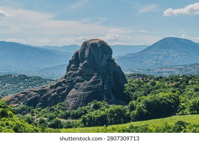 Horizontal landscape view of a massive monolithic dark grey rock formation surrounded by lush green forests and distant mountain ranges on a sunny summer day.