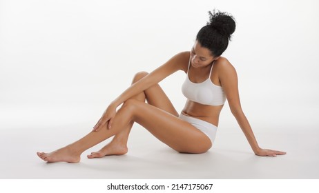 Horizontal Long Shot Of African American Woman In White Underwear Strokes Her Leg Sitting On The Floor On White Background | Smooth Legs And Unwanted Hair Removal Concept