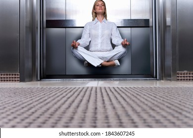 Horizontal low angle shot of a businesswoman meditating sitting cross-legged levitating in an open elevator in office with copy space.