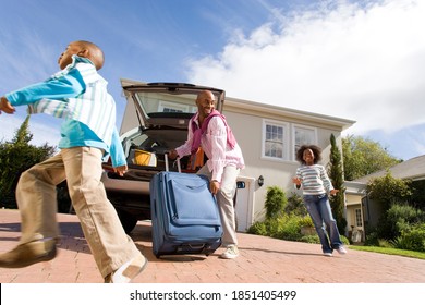 Horizontal low angle shot of two playful children running around by the father packing a suitcase into boot of a car on a sunny day.