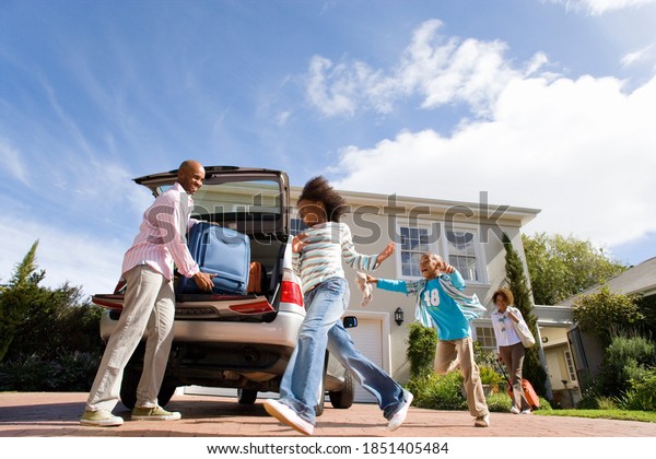 Horizontal low angle shot of two playful\
children running by the father packing a suitcase into a car with\
mother striding in the\
background.