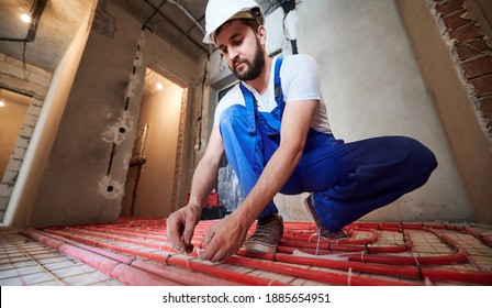 Horizontal, low angle view snapshot of young plumber wearing blue overalls and white helmet, tying up red tubes on the floor heating system in new unfinished apartment. Concept of home renovation.