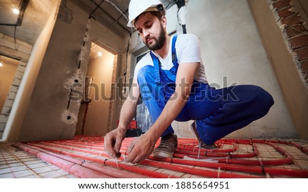 Horizontal, low angle view snapshot of young plumber wearing blue overalls and white helmet, tying up red tubes on the floor heating system in new unfinished apartment. Concept of home renovation.