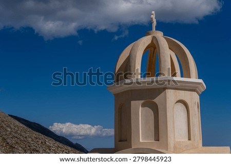 A horizontal low-angle shot of a classic stone church lantern structure with a cross, showcasing rustic textures against a vibrant blue sky with clouds in Santorini, Greece.