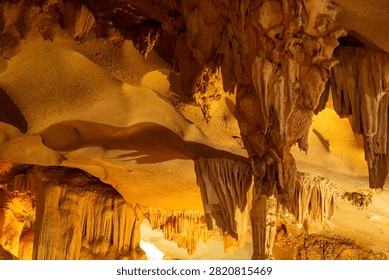 A horizontal low-angle shot features massive hanging stalactites, flowstone ceilings, and ancient karst structures brightly illuminated by golden artificial spotlights inside a subterranean cave at Ha Long Bay, Vietnam.