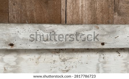 A horizontal macro photograph capturing a multi-layered weathered wooden structure featuring brown timber and painted white panels. The upper layer displays natural brown wood planks with vertical grain patterns and minor surface imperfections. A narrow horizontal wooden strip painted in distressed light gray stretches across the center, attached with dark rusty nails that show slight rust bleeding into the wood. The lower section reveals a heavily worn white painted wooden surface with visible paint peeling, scuffs, fine cracks, and natural weathering marks. This detailed composition offers a rich combination of colors, materials, and aged textures, highlighting traditional carpentry techniques and rustic architectural elements. Highly suitable for vintage background graphics, rustic wallpaper designs, architectural restoration projects, industrial grunge stock assets, wood texture overlays, and creative visual media. An authentic representation of vintage craftsmanship and natural wood decay.